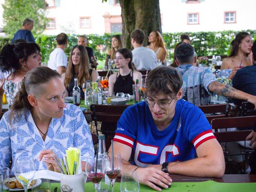 Zwei Kollegen sitzen unter einem großen Baum am Tisch im Biergarten, im Hintergrund weitere Gäste.